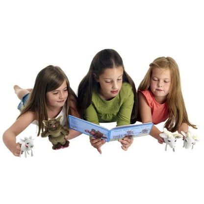 Three young girls lie on their stomachs while reading a book together, surrounded by small toy figures and finger puppets from The Puppet Company Finger Puppet Story Set Three Billy Goats Gruff. The scene appears relaxed and engaging, suggesting they are enjoying their storytelling and playtime.