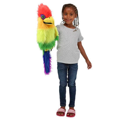 A child with braided hair smiles and holds The Puppet Company Rainbow Bird Hand Puppet. This vibrant bird puppet, adorned with red, green, yellow, and purple feathers, features a squeaker in its mouth. Dressed casually in a striped t-shirt, jeans, and red sandals, the child enjoys playing as the puppet's beak opens as if speaking.