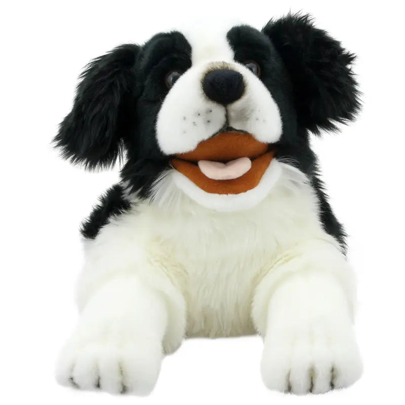 The Border Collie Hand Puppet - Playful Puppies features black and white fur, floppy ears, an open mouth with a pink tongue and white teeth, and lies down with its front paws stretched forward.
