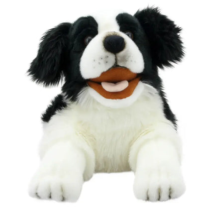 The Border Collie Hand Puppet - Playful Puppies features black and white fur, floppy ears, an open mouth with a pink tongue and white teeth, and lies down with its front paws stretched forward.