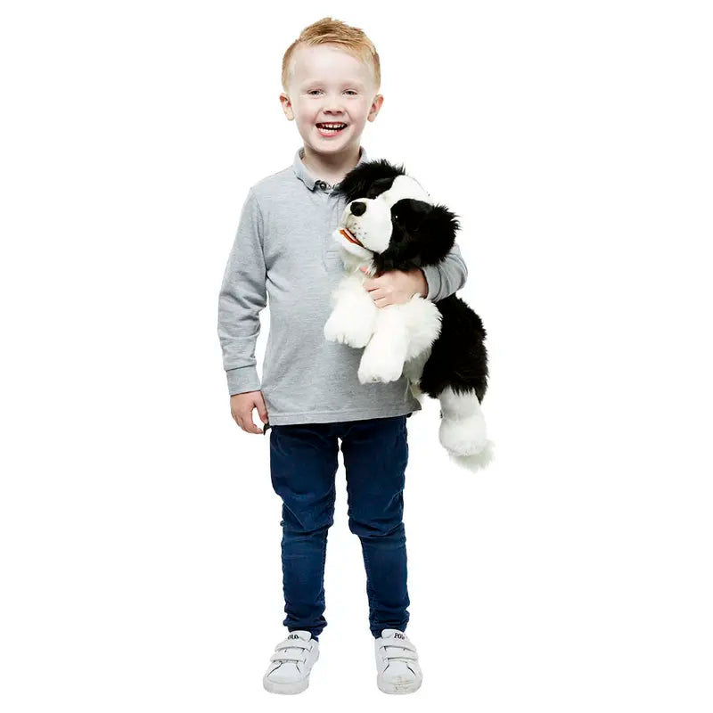 A smiling young boy with light hair, in a grey long-sleeve shirt and blue pants, holds the Border Collie Hand Puppet - Playful Puppies against a plain white background.