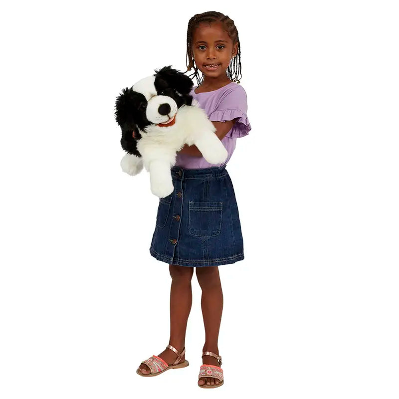 A young girl with braids, dressed in a lavender shirt and denim skirt, smiles while holding the Border Collie Hand Puppet - Playful Puppies.