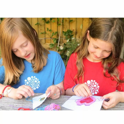 Two girls are making crafts with a Buttonbag Bumper Cross Stitch Kit at a table outdoors.