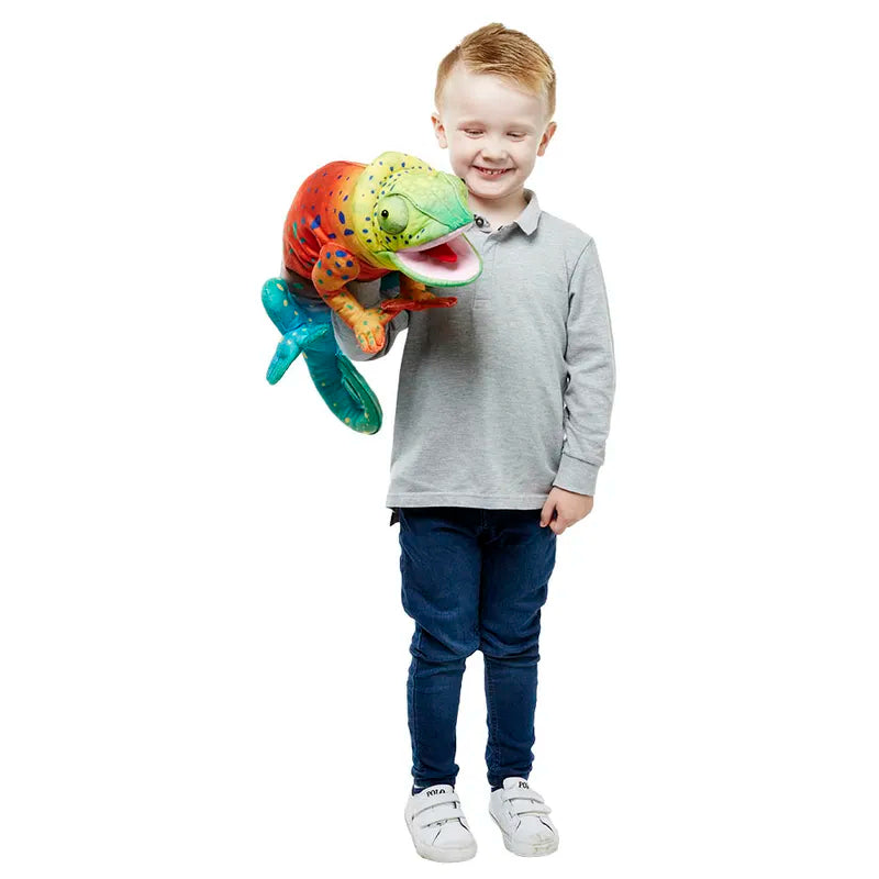 A young child with short hair is standing and smiling while holding The Puppet Company Large Creature Chameleon. The child is wearing a grey long-sleeve shirt, blue jeans, and white sneakers. This children's toy fish is bright with patterns and has realistic movements that make it look friendly.