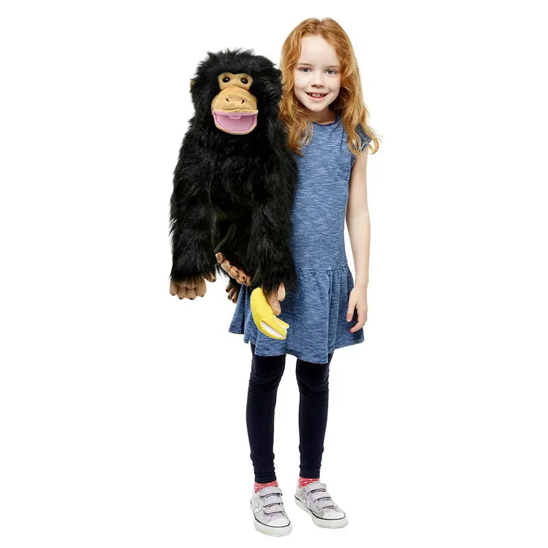 A smiling young girl in a blue dress holds The Puppet Company Medium Puppet Primate Chimp, a mouth-moving hand puppet with soft fur and a banana in its hand, against a plain white background.