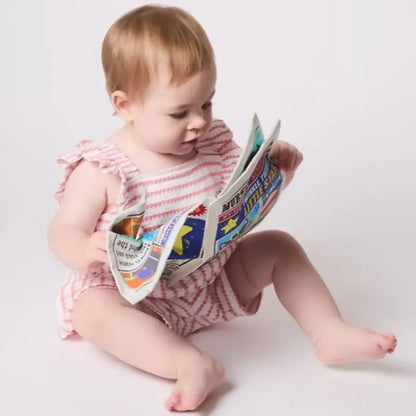 A baby in a pink and white striped romper sits on a plain white floor, holding and looking at the Crinkly Cloth Book Nursery Rhymes 1.