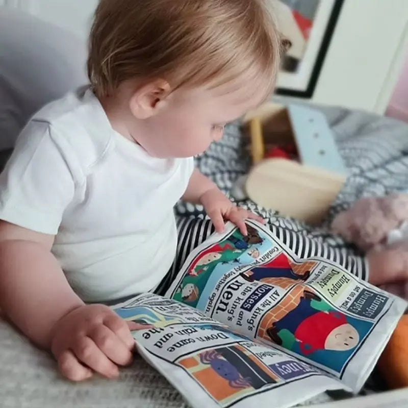 A toddler in a white shirt sits on a bed, exploring the Crinkly Cloth Book Nursery Rhymes 1. Toys and a stuffed animal are in the background, creating a playful nursery scene perfect for sharing rhymes.