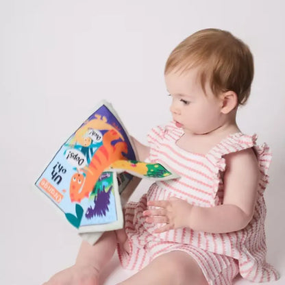 A baby in a pink and white striped romper sits and focuses on the Crinkly Cloth Book Rainbow Dinosaurs, captivated by its colorful dinosaur illustrations and sensory features against a plain light background.