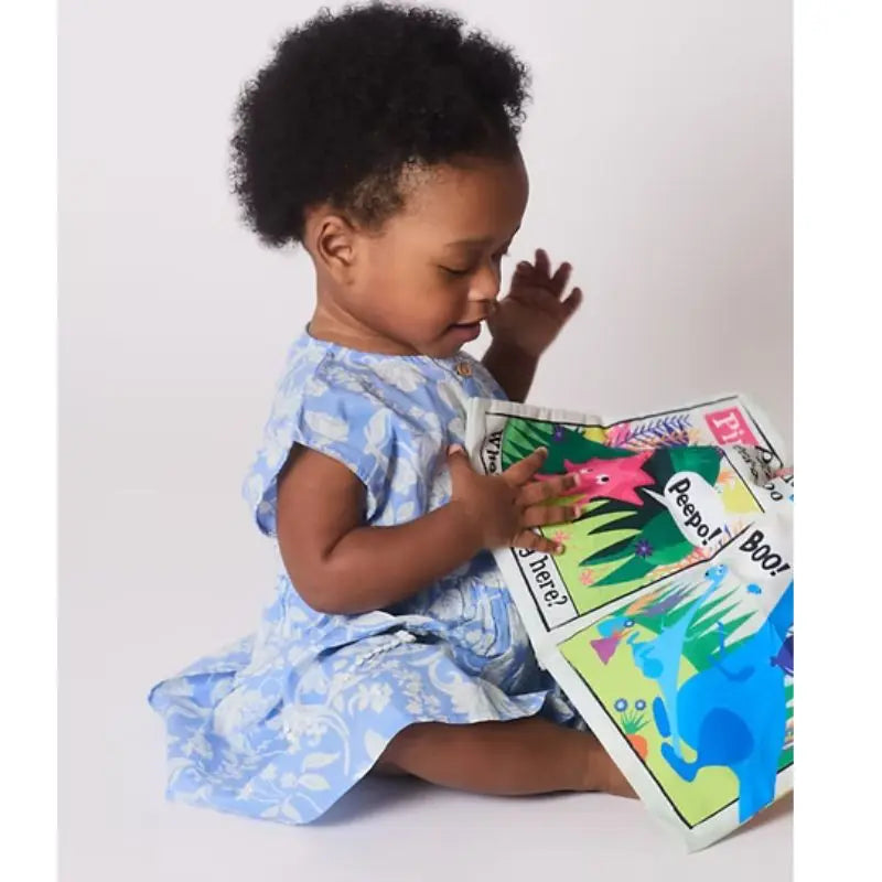 A toddler in a blue floral dress sits on the floor, captivated by the Crinkly Cloth Book Rainbow Dinosaurs—a sensory-rich baby book with pop-up features and vibrant illustrations, perfect for little hands.