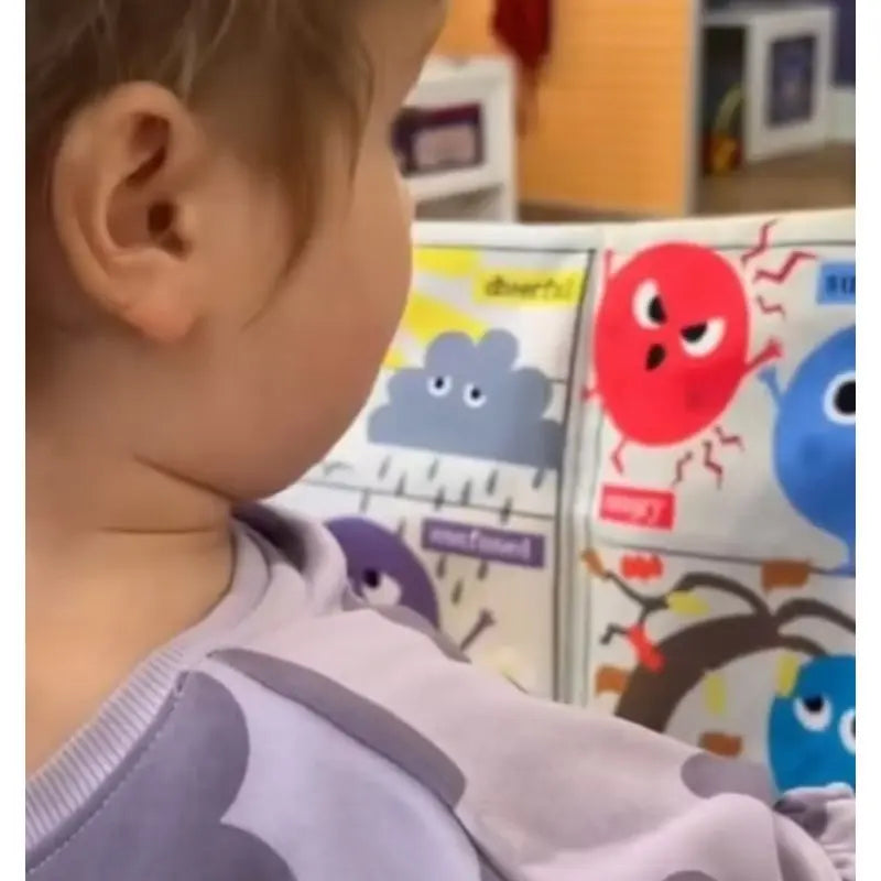 A young child wearing a light purple shirt explores the Crinkly Cloth Book Rainbow of Emotions, featuring colorful cartoon clouds and expressive characters—ideal as a baby emotions book or a thoughtful newborn gift.