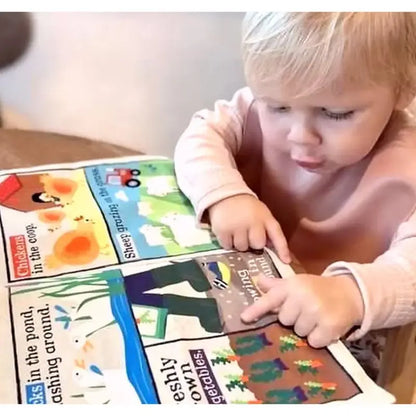 A young blonde-haired child in a pink long-sleeve shirt sits at a table, pointing to colorful animals inside the open Crinkly Cloth Book Farm Animals.