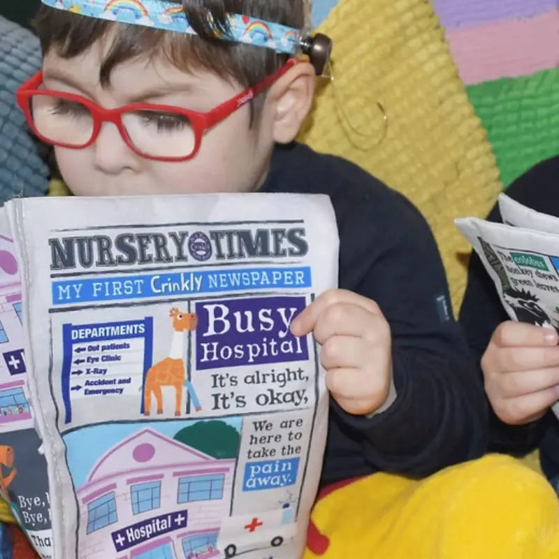 A young child with red glasses reads the colorful, crinkly cloth book "Crinkly Cloth Book Hospital" while sitting on a textured blanket—an ideal baby gift. Another child is partially visible, also holding this playful toy newspaper.