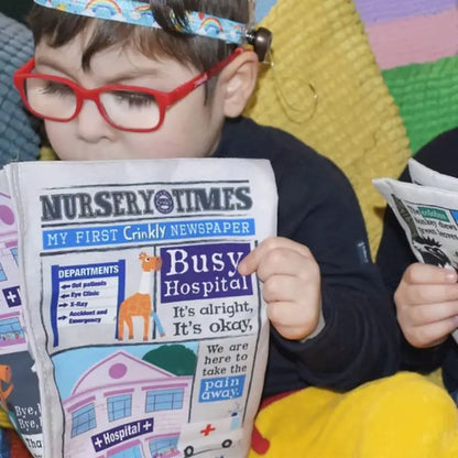 A young child with red glasses reads the colorful, crinkly cloth book "Crinkly Cloth Book Hospital" while sitting on a textured blanket—an ideal baby gift. Another child is partially visible, also holding this playful toy newspaper.