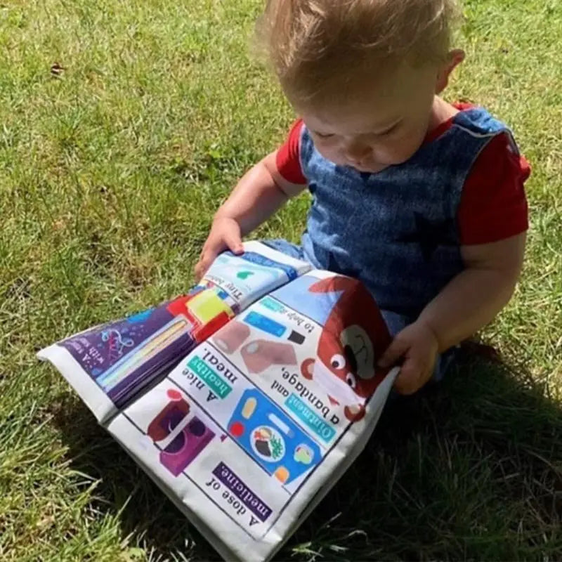 A young child sits on grass, focused on the Crinkly Cloth Book Hospital open on their lap. Filled with bright illustrations and playful characters, it's a perfect baby gift. Sunlight enhances the cheerful scene.