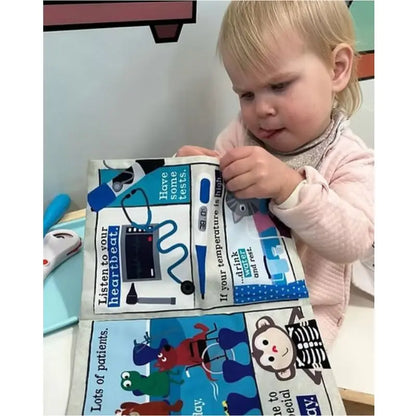 A young child in a pink sweater explores the Crinkly Cloth Book Hospital, an interactive, colorful cloth book featuring medical illustrations and text about heartbeats and stethoscopes.