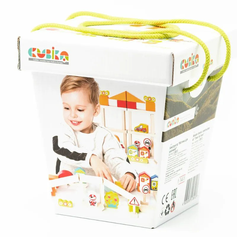 The image shows packaging for a children's toy set by "Cubika." The box is white with colorful illustrations and images of the "Cubika ECO Wooden Blocks Construction City" toy set, featuring toddler wooden blocks and a smiling child. Green rope handles on the top make it easy to carry, while the packaging promotes imaginative play. Safe water-based paints are highlighted in the product information printed on the sides.