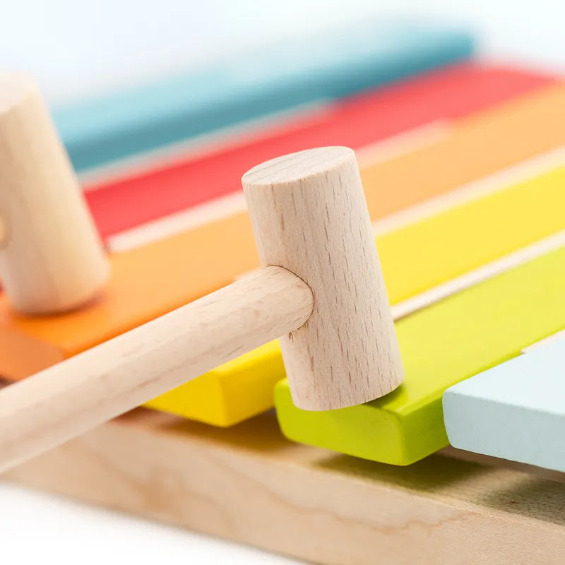 The Cubika Wooden Xylophone, a vibrant toddler instrument, features colorful rainbow bars in blue, red, orange, yellow, and green. A wooden mallet rests atop this delightful tool that invites children to explore music.