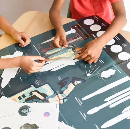 Two children place stickers on the Poppik Discovery Sticker Poster – Astronomy, a colorful solar system chart with rockets, spacecraft, and labeled diagrams, spread out on a wooden table.