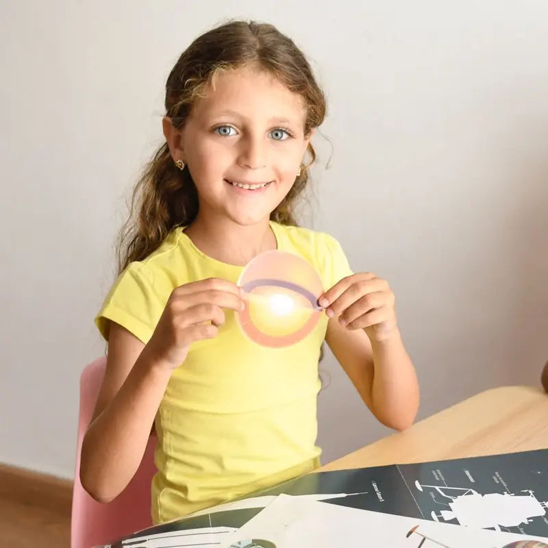 A young girl with long curly hair and blue eyes, wearing a yellow t-shirt, sits at a table smiling and holding the Poppik Discovery Sticker Poster – Astronomy. Papers, charts, and a colorful astronomy poster are spread out on the table.