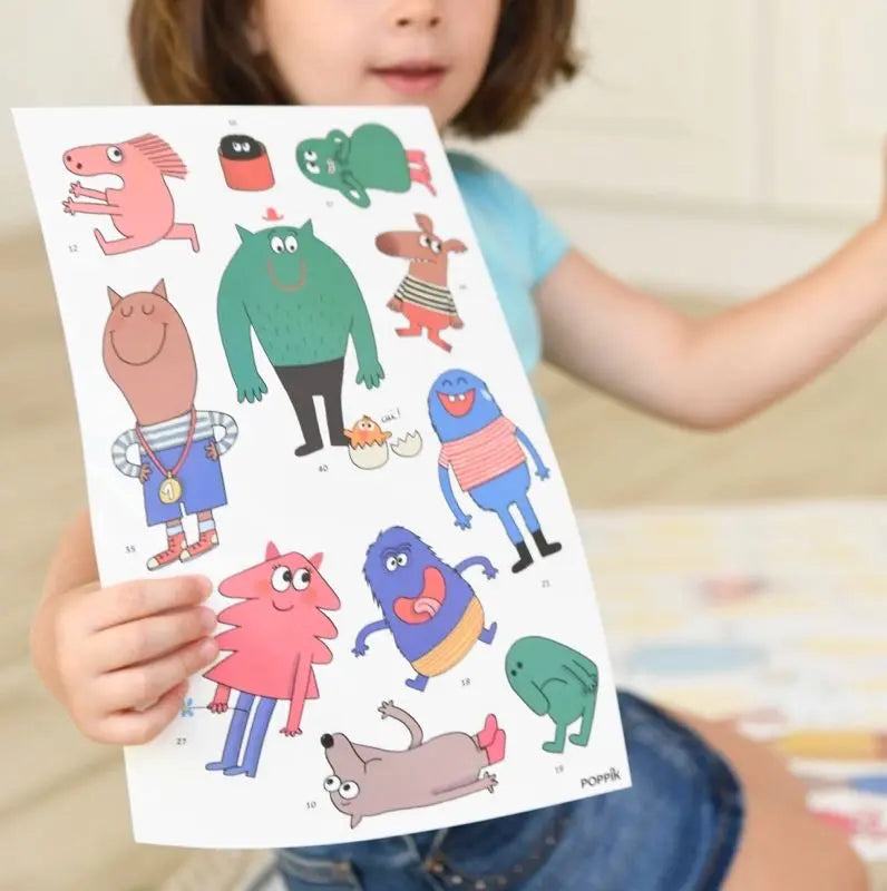 A child sits on the floor beside the Poppik Discovery Sticker Poster – Emotion, holding up a sheet of colorful cartoon monster stickers in various shapes, sizes, and colors.