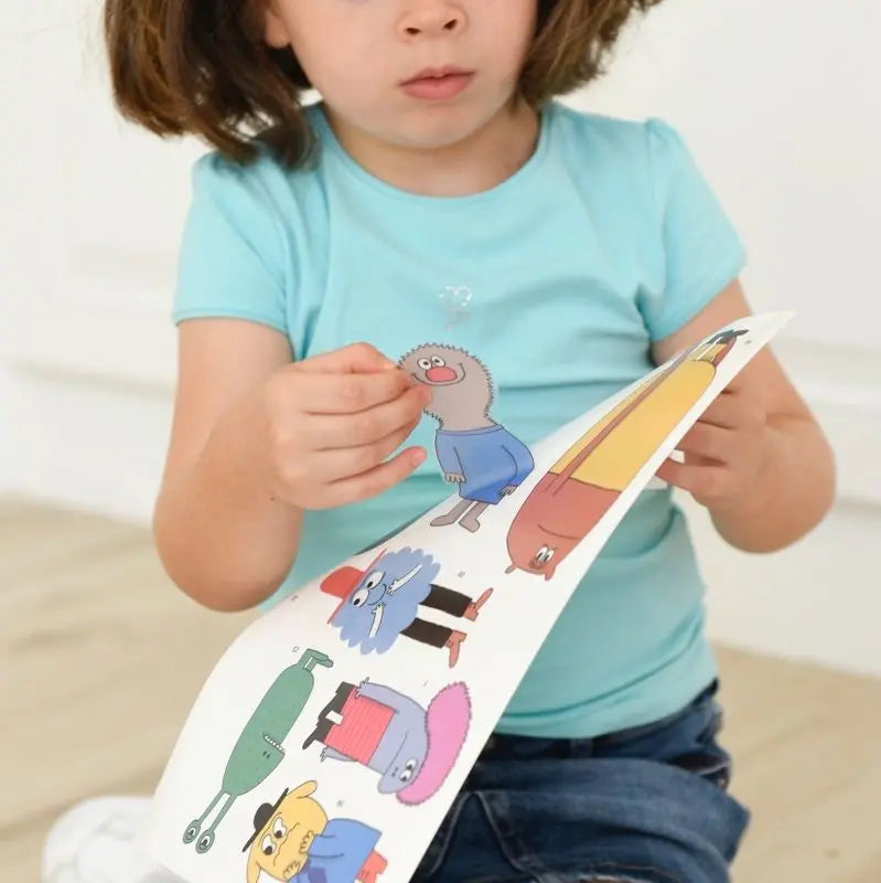 A young child in a light blue shirt peels and holds a sheet of cartoon stickers from the Poppik Discovery Sticker Poster – Emotion, looking down with focus—perhaps preparing to decorate an educational poster.