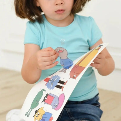 A young child in a light blue shirt peels and holds a sheet of cartoon stickers from the Poppik Discovery Sticker Poster – Emotion, looking down with focus—perhaps preparing to decorate an educational poster.