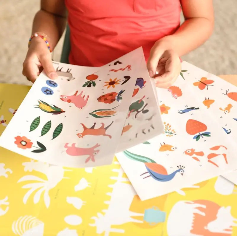 A child holds two sheets from the Poppik Discovery Sticker Poster – Farm, featuring colorful animal and nature-themed stickers, with yellow paper and extra farm animal stickers visible—an engaging activity for children.