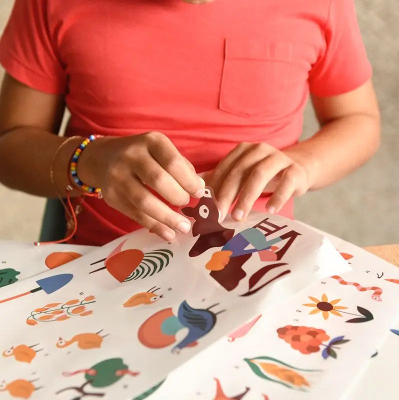 A person in a red shirt peels an animal sticker from the Poppik Discovery Sticker Poster – Farm, which features illustrated animals, plants, and objects. Only their torso and hands are visible.