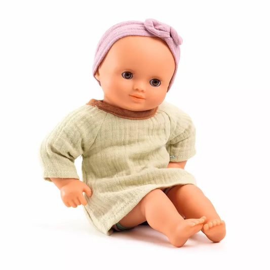 A Djeco Baby Pistache baby doll sitting on the ground wearing a pink headband.