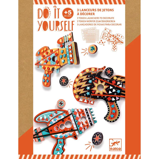 The Djeco Mosaics & Stickers Kosmik comes in an orange and white package designed for a DIY craft kit suitable for kids aged 5 and up. This creative kit includes materials to assemble and decorate three vibrant token launchers, featuring mosaic stickers with assorted patterns and designs. The packaging text is presented in multiple languages.