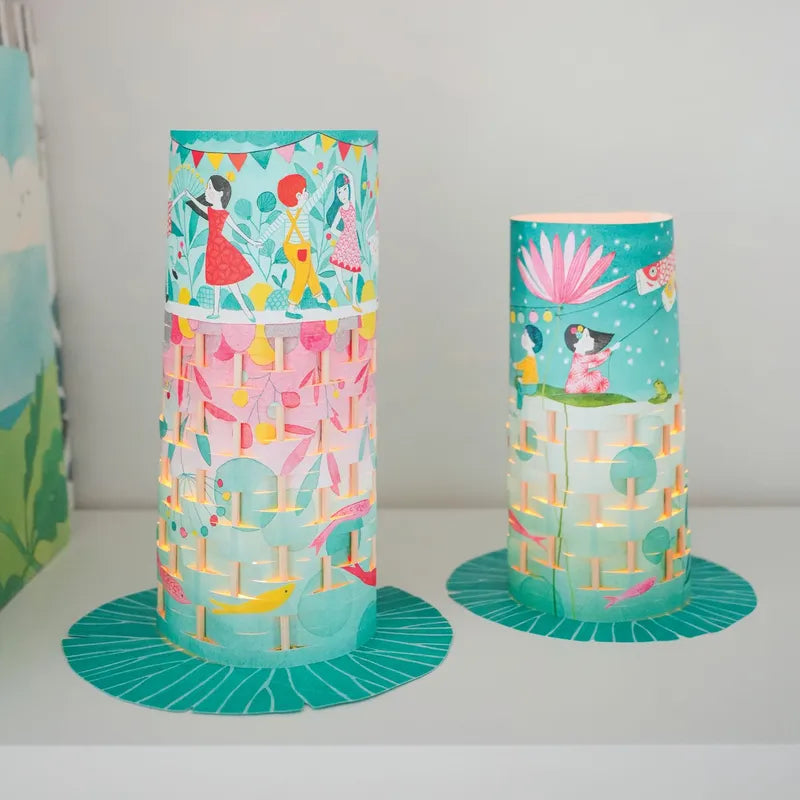 Two cylindrical Djeco Create Fireflies Dance paper lanterns are beautifully displayed on a white surface. These colorful lanterns feature intricate illustrations of people, animals, and nature scenes in vibrant hues of blue, pink, and green. Their unique cut-out pattern and delicate paper weaving add charm, allowing light to shine gracefully through.