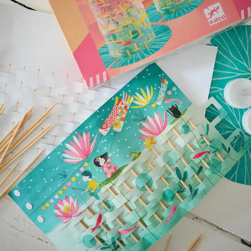 The Djeco Create Fireflies Dance kit features paper templates with playful illustrations of fish, flowers, and water lilies, bamboo sticks, and a circular blue mat. One paper weaving is partially assembled, showing a pond theme reminiscent of delicate paper lanterns.