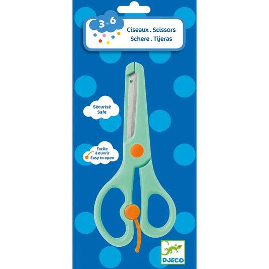 A pair of Djeco Accessories For Little Ones Scissors, featuring a green, child-safe design with an orange button on the handle for kids ages 3-6, is displayed on a blue polka-dotted cardboard backing. The packaging includes the words "Ciseaux, Scissors, Schere, Tijeras" and prominently shows the "Djeco" logo at the bottom right.