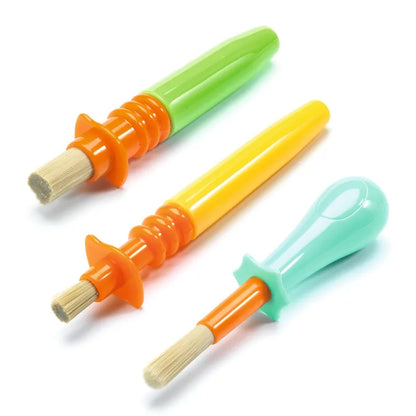 The Djeco Accessories For Little Ones 3 Ingenious Paintbrushes are shown with colorful handles perfect for little hands. One brush features a green handle with an orange base, another sports a yellow handle with an orange base, and the third has a light blue handle paired with an aqua blue base. Each brush has short, bristly tips and includes a no-roll design to prevent them from rolling away.