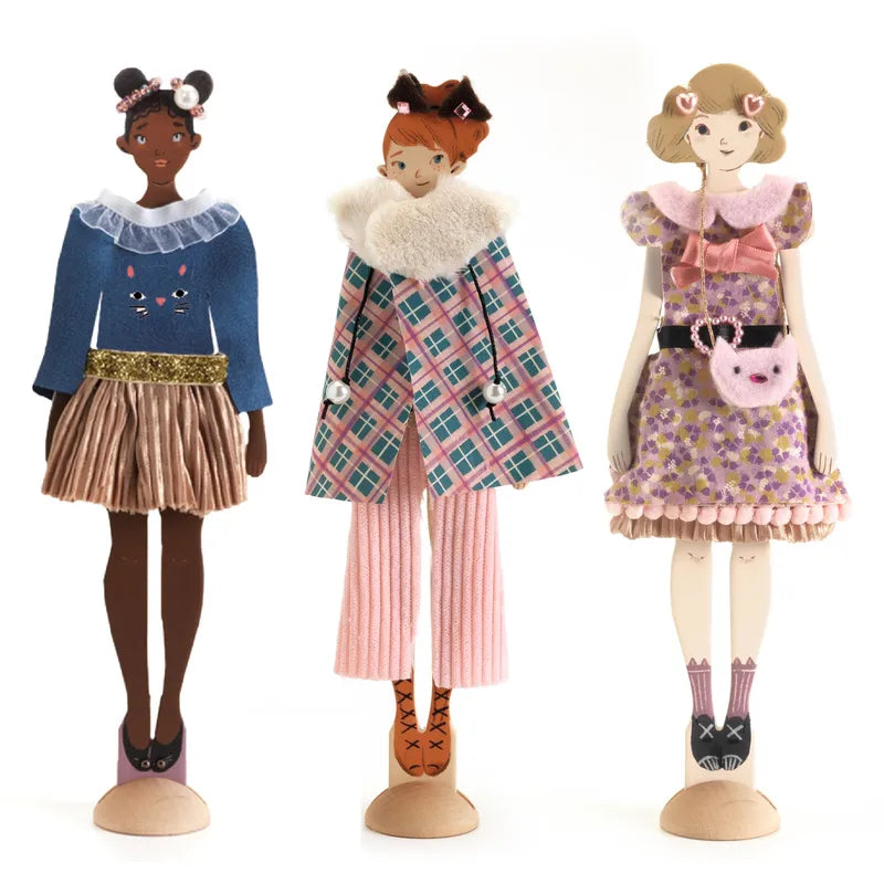 Displayed are three chic doll figures, created by young fashion enthusiasts using the Djeco Workshops Fashion by Lucille set. Each doll sports distinctive attire with animal-themed accessories such as cat ears and playful prints. The outfits, in hues of blue, pink, and brown, showcase an imaginative style—all crafted without any sewing.