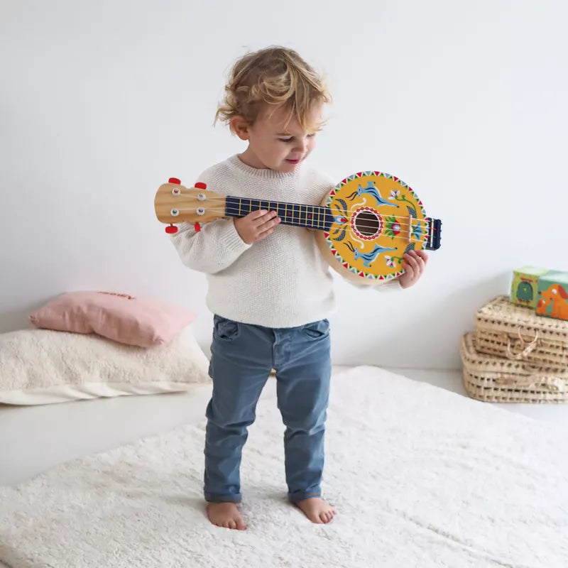 A little boy standing on a bed holding a Djeco Animambo Banjo.