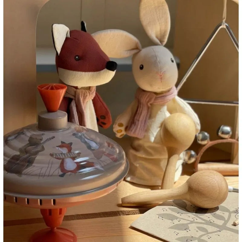 A cozy scene with a plush fox and rabbit in scarves is set among wooden toys, highlighted by a charming tin toy. Adding to the warmth and sunlight is the Large Spinning Top Musicians with animal illustrations and a triangle instrument.