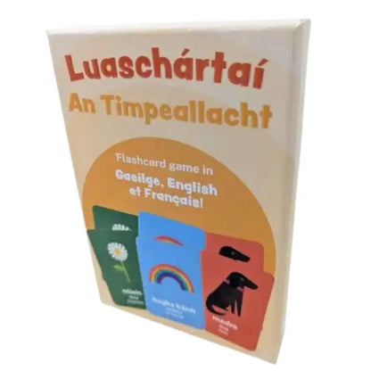 Luaschártaí - An Timpeallacht" is a box of trilingual flashcards for language learning, featuring Irish, English, and French. The front cards show colorful images of a daisy, rainbow, and dog.