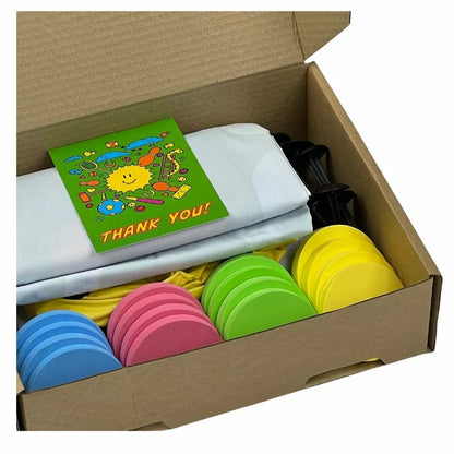 Open cardboard box containing a white folded t-shirt with a colorful "thank you" card on top, and stacked colorful silicone travel bottles in pink, green, and yellow alongside a Giant Ludo 1.5m x 1.5m