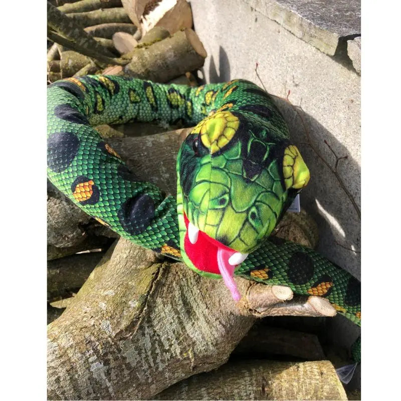 The Puppet Company Large Creature Snake—a realistic-looking green plush with black and yellow markings and a red forked tongue—rests on cut logs by a concrete wall, inviting imaginative nature-inspired play.