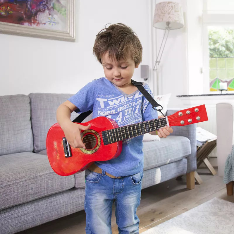 A young boy playing a New Classic Toys Red Guitar in a living room.