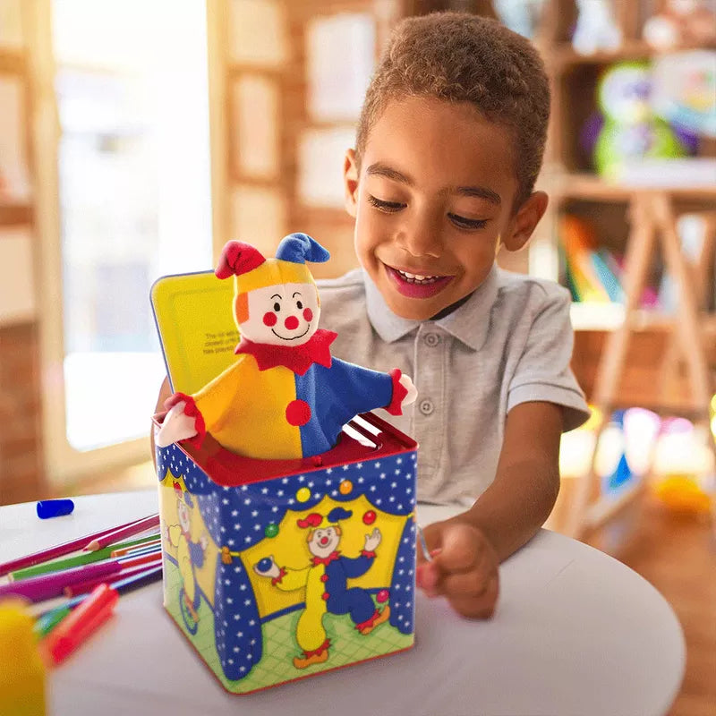 A young boy smiling at a colorful Jester Jack In Box toy in a bright, art-filled room. The toy for toddlers features a clown popping out of a blue box with yellow stars.