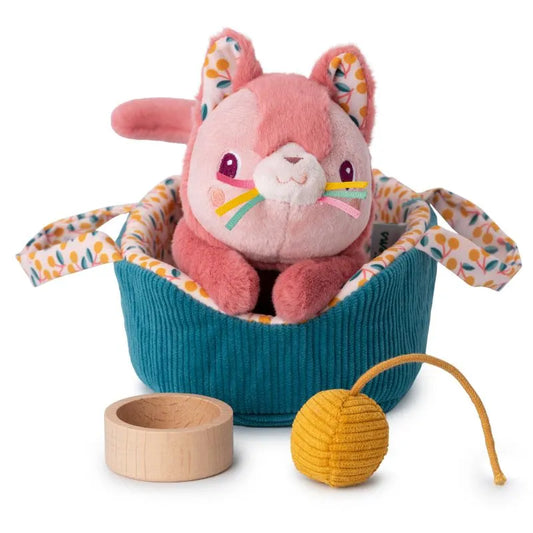 The Lilliputiens "I'm Exploring Jeanne Cat With Basket" is an adorable plush pink cat with rainbow whiskers nestled in a teal basket, making it a perfect baby gift, along with a small wooden bowl and a yellow ball for delightful toy adventures.