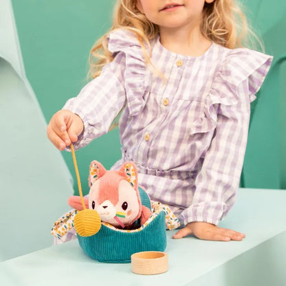 In a soft green background, a child in a gingham dress delights with the Lilliputiens I'm Exploring Jeanne Cat With Basket. The plush toy, pink with ears, sits in the teal basket as the child grips a string holding a small yellow object.