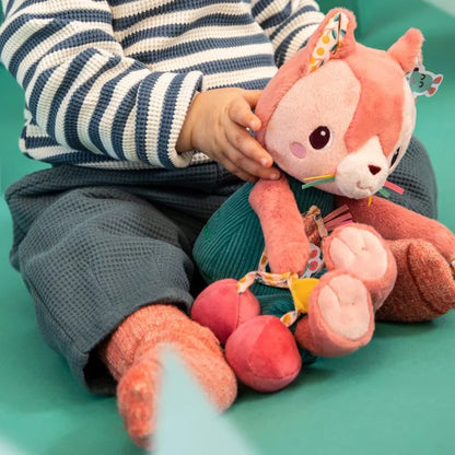 A baby, dressed in a striped sweater and textured pants, sits on a teal surface holding the Lilliputiens I'm Discovering Jeanne Multi-Activity Cat. This plush toy, with its embroidered details and friendly expression, is perfect as a baby's first toy and an ideal gift.