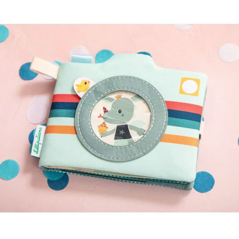 The Lilliputiens I'm Discovering Joe Photo Album is a camera-shaped, pastel-colored item with a cute character visible through a circular window. It features colorful stripes, a fabric tag, and is suitable for infants 9 months+. The scattered confetti backdrop enhances its playful design.