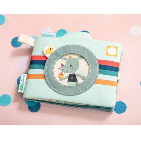 The Lilliputiens I'm Discovering Joe Photo Album is a camera-shaped, pastel-colored item with a cute character visible through a circular window. It features colorful stripes, a fabric tag, and is suitable for infants 9 months+. The scattered confetti backdrop enhances its playful design.