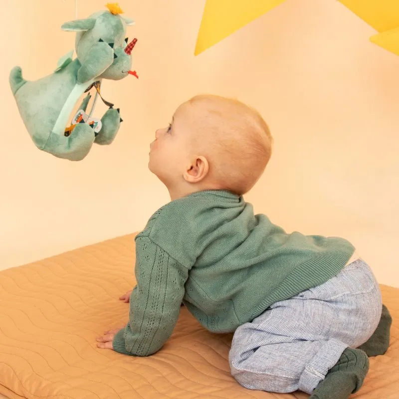 A baby in a green sweater and light blue pants is crawling on a quilted surface, eagerly reaching for their Lilliputiens I'm Discovering Joe Peekaboo Mirror—a green stuffed dragon hanging above. The background is soft and warm-toned with a yellow star shape.