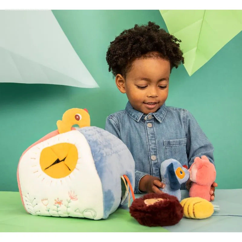 A curly-haired child in a denim shirt explores plush toys against a vibrant green background, including the colorful "Lilliputiens I'm Discovering Jules' House And 4 Hidden Rattles" set—a wonderful choice for a baby's first toy or an ideal baby gift featuring a soft house, bird, and fox.