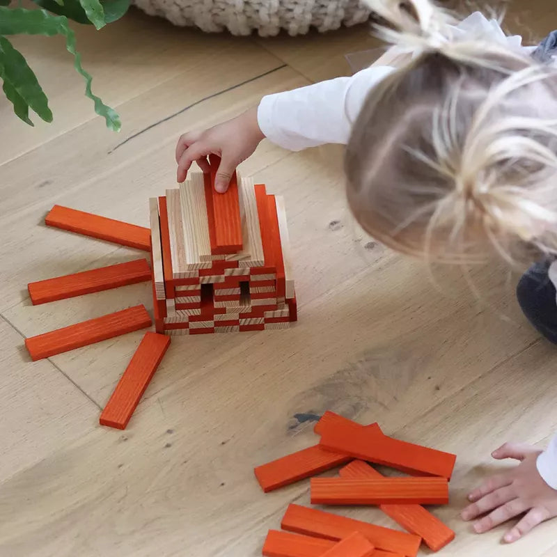 A child engages in a construction game with KAPLA® 40 Coloured Planks Orange, carefully adding a piece to a creatively stacked structure on a wooden floor.